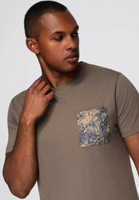 Olivově zelené tričko s krátkým rukávem se čtvercovou kapsou v maskáčovém vzoru, obsahující odstíny zelené, béžové a modré. Měkká textura a klasický střih.