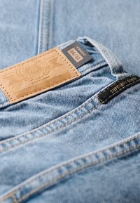Vaqueros de mezclilla azul claro con un parche de cuero marrón claro que dice "SUPERDRY" y la talla "26/32". Tienen un corte recto y detalles de costura visibles.