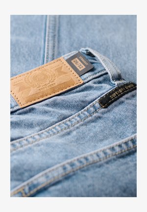Lichtblauwe spijkerbroek van denim met een tan leren patch met het label "SUPERDRY" en maat "26/32." Beschikt over een rechte snit en zichtbare stikseldetails.