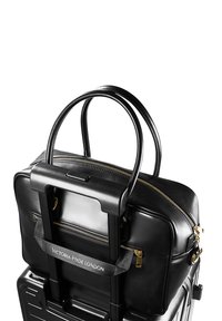 Schwarze Leder-Handtasche an einem Griff eines schwarzen Rollkoffers befestigt, mit goldenen Reißverschluss-Details und einem Markenriemen mit der Aufschrift "Victoria Hyde London."