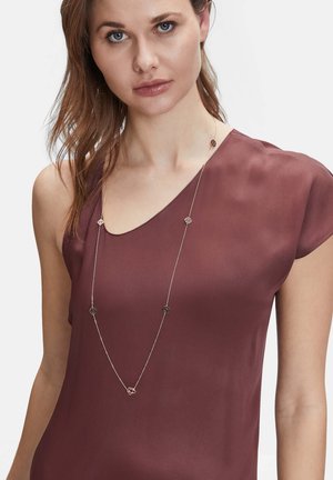 Bordeauxrotbluse mit weicher Textur und kurzen Ärmeln, kombiniert mit einer silbernen Kettenhalskette, die geometrische Akzente und kreisförmige Details aufweist.