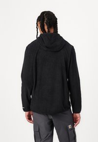 Felpa nera in pile con finitura testurizzata, tasca a marsupio e cappuccio. Maniche lunghe con polsini elasticizzati. Indossata sopra pantaloni cargo grigi.