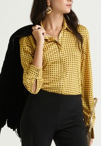 Camicia gialla con bottoni, a pois neri, maniche lunghe con risvolti; abbinata a pantaloni neri a vita alta e giacca nera.