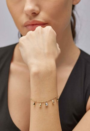 Bracciale in oro con cinque accenti a forma di goccia e un gemma trasparente, esposto su un polso contro uno sfondo di abbigliamento scuro.