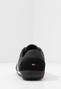 Sneaker nera con pannello in suede sul tallone, caratterizzata da strisce bianche e rosse lungo il lato e una parte posteriore con il marchio 'HILFIGER' in rilievo.