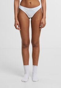 Valge pitsäärtega bikiinialuspesu, millel on sile viimistlus ja lainelised servad, koos valgete pahkluusokkidega, millel on volangiga ülemine osa.