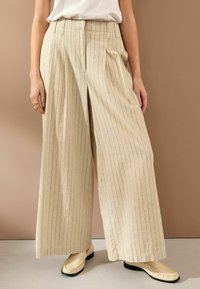 Beige weite Hose mit senkrechten blauen Streifen, aus Leinenstoff, mit vorderen Falten und hoher Taille, kombiniert mit cremefarbenen Slippern.