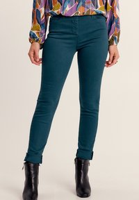 Jeans skinny turchesi con vita media, texture liscia e orli alla caviglia abbinati a stivaletti neri con tacco.