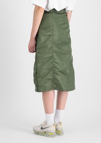 Gonna verde di lunghezza media realizzata in tessuto lucido e arricciato. Indossata con calzini bianchi e scarpe da ginnastica, con cuciture visibili e una superficie liscia.