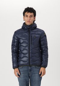 Giubbotto in piumino blu navy con design trapuntato, zip frontale e cappuccio. Presenta una piccola tasca sul petto e accenti verdi a contrasto.