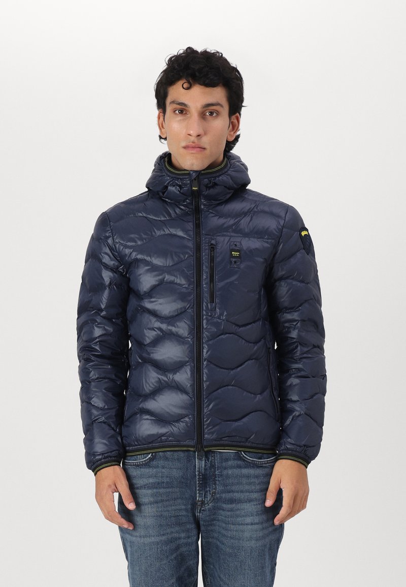 Giubbotto in piumino blu navy con design trapuntato, zip frontale e cappuccio. Presenta una piccola tasca sul petto e accenti verdi a contrasto.