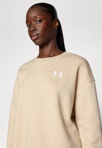 Beige sweatshirt med teksturert overflate, med rund hals, senkede skuldre og hvitt brodert logo på brystet.