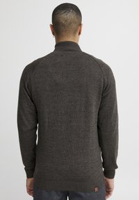 Suéter de punto gris oscuro con un cuello alto, puños y dobladillo acanalados, que presenta un pequeño parche de logo en la parte baja de la espalda. Corte clásico y material texturizado.