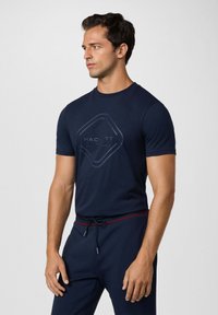 Mörkblå bomullströja med korta ärmar, med en diamantformad HACKETT-logotyp i en mörkare nyans på framsidan. Enkel design, inga mönster.