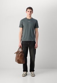 Grå kortermet T-skjorte, mørkeblå jeans, grønne og beige joggesko, brun lær duffelbag med doble håndtak og hardware-detaljer.