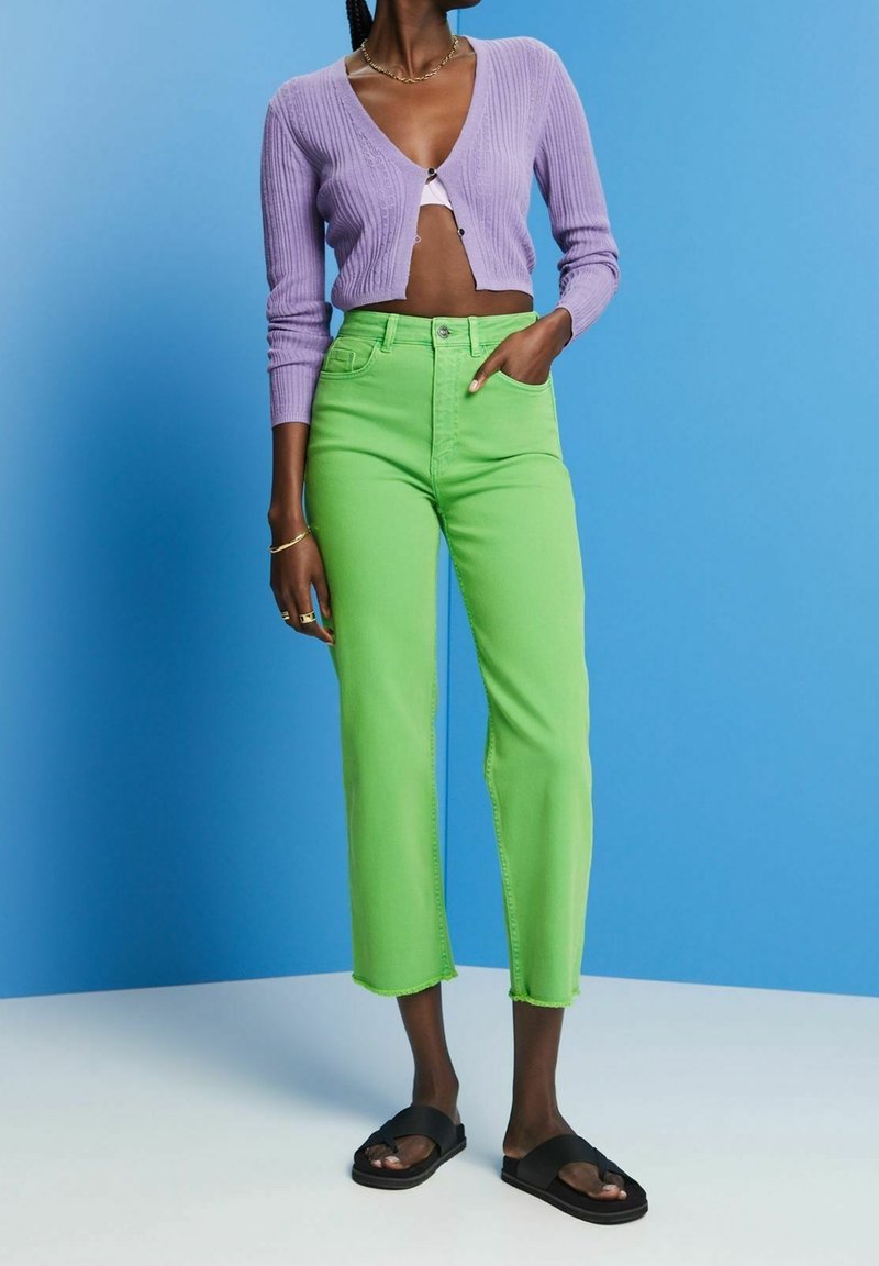 Femme portant un cardigan court lavande, un bralette blanc, un jean vert citron taille haute et court, ainsi que des sandales noires à enfiler, sur fond bleu.
