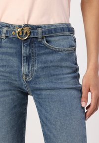 Augstais jostasvietas zila džinsu bikses ar nolietotu finišu, kas aprīkotas ar zelta jostu sprādzi ar dekoratīviem putniem un klasisku piecu kabatu dizainu.