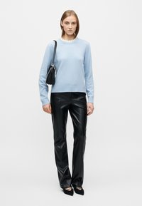 Ανοιχτό μπλε πουλόβερ με ριμπ λαιμόκοψη, σε συνδυασμό με μαύρο γυαλιστερό παντελόνι από συνθετικό δέρμα. Το μοντέλο κρατά μια μικρή μαύρη τσάντα με λουράκι αλυσίδα.