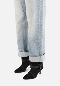 Must musta anke saapad terava varba, millel on kootud detailid ja väike, virnastatud konts, koos rullitud heledate denim teksadega.