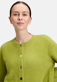Femme avec les cheveux tirés en arrière, portant un cardigan en mohair vert lime avec quatre boutons ronds brillants, sur un fond blanc.