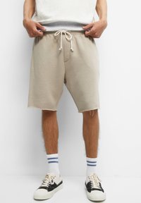 Homme portant un short beige à cordon de serrage, des chaussettes blanches à rayures bleues et des baskets noir et blanc, debout sur un fond blanc.