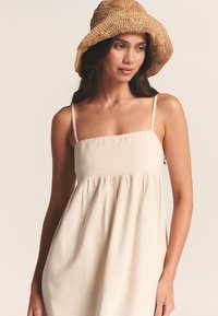 Smėlio spalvos lininė suknelė su kvadratine iškirpte, plonomis petnešėlėmis ir pūsta suknele. Derinama su austu, apvalia kraštine šiaudiniu skrybėle.