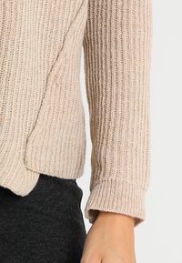 Beige neulepaita, jossa on resorimainen tekstuuri, pitkät hihat, joiden hihansuut on käännetty, ja löysä istuvuus helmassa.