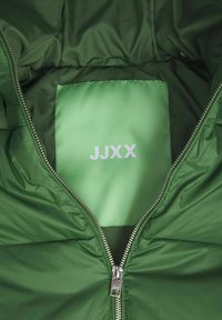 Grön vadderad jacka med en slät yta, silverfärgad dragkedja och en fyrkantig etikett inuti tryckt med "JJXX" i vitt.