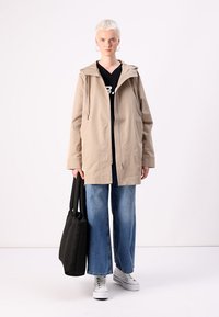 Casaco leve bege com capuz, com fecho à frente, dois bolsos laterais, combinado com uma t-shirt preta, calças de ganga largas azuis e ténis brancos.