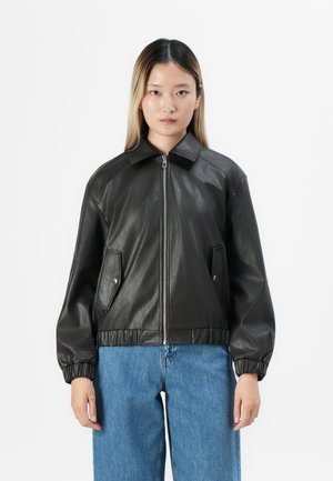 Ungdom med rett hår som har på seg en svart lærjakke med glidelås og elastiske mansjetter, samt blå jeans, stående mot en hvit bakgrunn.