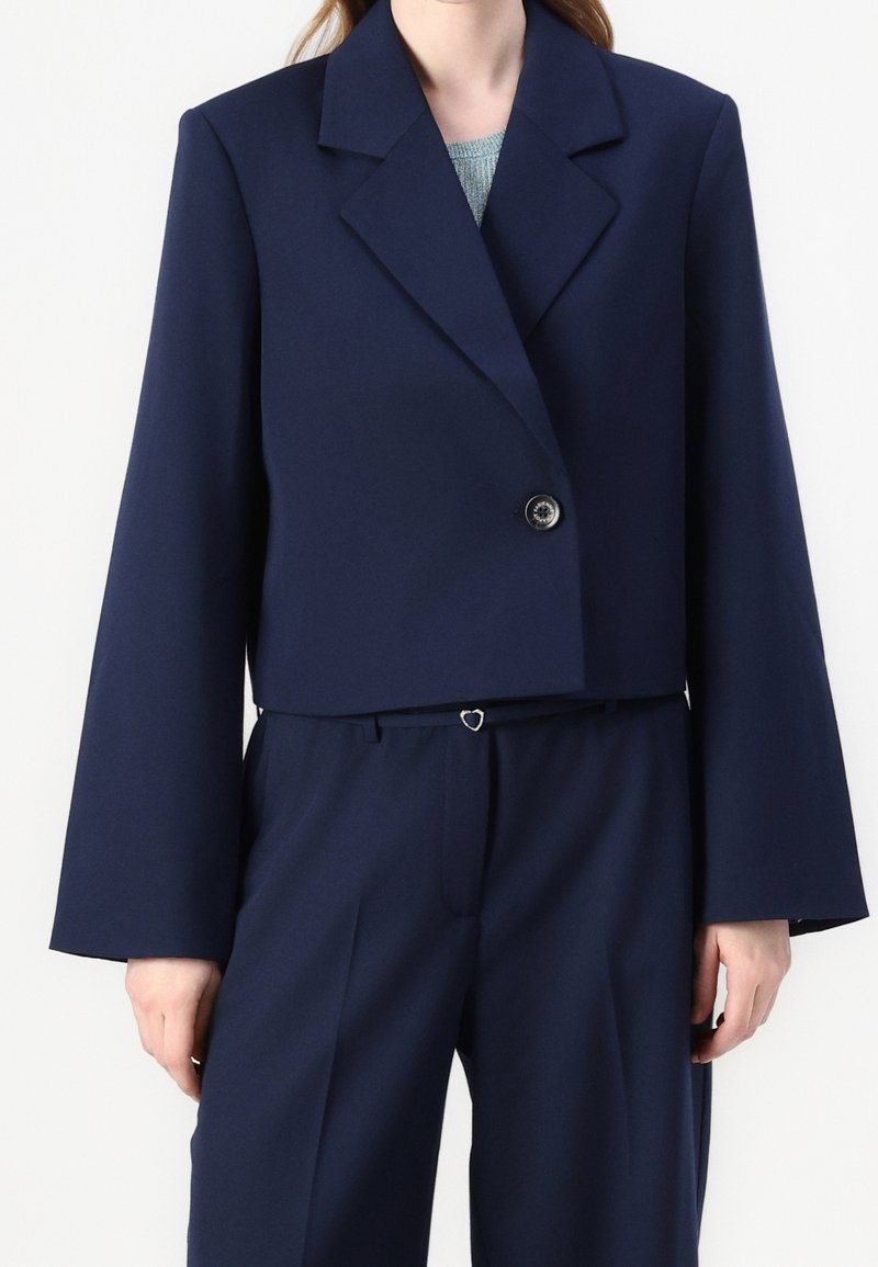 Femme portant un blazer court bleu marine avec un seul bouton et un pantalon taille haute assorti avec une petite boucle de ceinture en forme de cœur.
