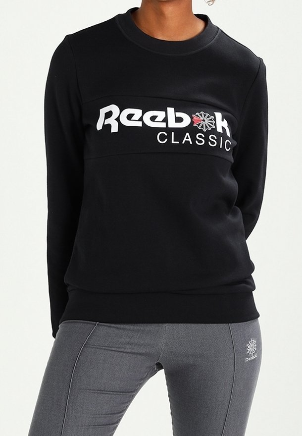 Personne portant un sweat-shirt noir Reebok Classic à manches longues et un legging gris Reebok, debout les mains derrière le dos.
