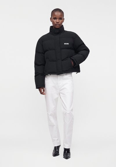 Personne portant une doudoune noire Moschino, un jean blanc avec des patchs en forme de cœur aux genoux et des bottines noires, se tenant devant un fond blanc.
