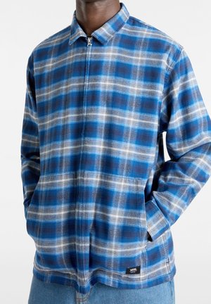 Mężczyzna ubrany w niebiesko-szarą kurtkę w kratę na zamek z kołnierzem i bocznymi kieszeniami, noszący jasnoniebieskie dżinsy.