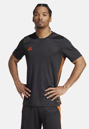 Mężczyzna z zaplecionymi włosami, ubrany w czarno-pomarańczowy strój sportowy Adidas, stoi z rękami na biodrach, zwrócony przodem, o neutralnym wyrazie twarzy.