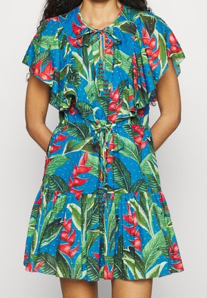 Mujer que lleva un vestido azul de manga corta con flores tropicales rojas, hojas verdes, mangas con volantes, borlas de cuentas y un cinturón en la cintura.