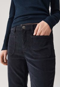 Vrouw in een donkerblauw geribbeld T-shirt met lange mouwen en zwarte corduroy broek met voorzakken, hand rustend in de rechterzak.