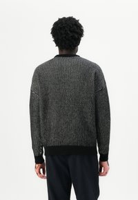 Maglione lavorato a maglia in bianco e nero con un motivo testurizzato, maniche lunghe, collo rotondo e orlo e polsini a costine. Osservato da dietro.