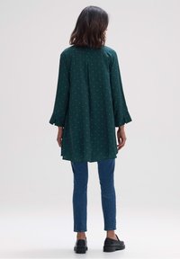 Tunic verde con una vestibilità lunga e fluttuante, caratterizzata da piccoli punti bianchi. Il materiale sembra leggero, con maniche lunghe e spacchi laterali. Abbinata a jeans.