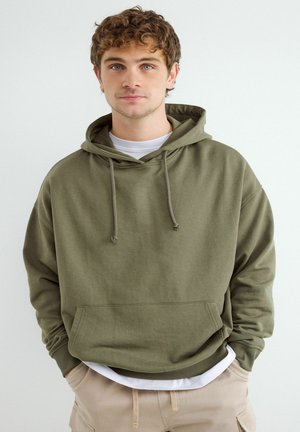 Junger Mann mit lockigem Haar, der einen olivgrünen Hoodie und beige Hosen trägt, steht mit den Händen in den Taschen vor einem schlichten Hintergrund.