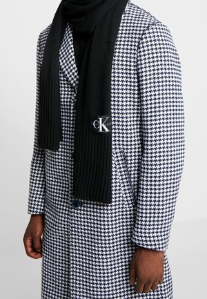 Homme portant une écharpe noire en maille avec le logo Calvin Klein blanc sur un manteau à motif pied-de-poule noir et blanc.
