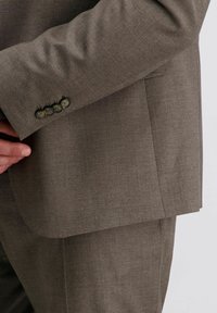 Rechter Arm passt den Manschettenknopf einer braunen Sakkojacke mit vier Knöpfen in der Nähe des Handgelenks an.