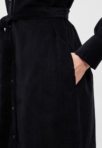 Černé šaty z manšestru s páskem v pase, zapínáním na knoflíky a bočními kapsami; mají vertikální žebrování pro texturu.