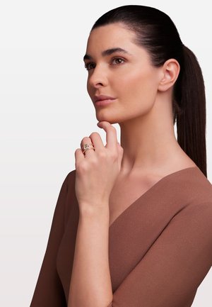 Gold- und Silberring mit verschlungenen Bändern und einem zentralen Stein. Auf einer Hand mit hellbrauner Haut, vor einem neutralen Hintergrund.