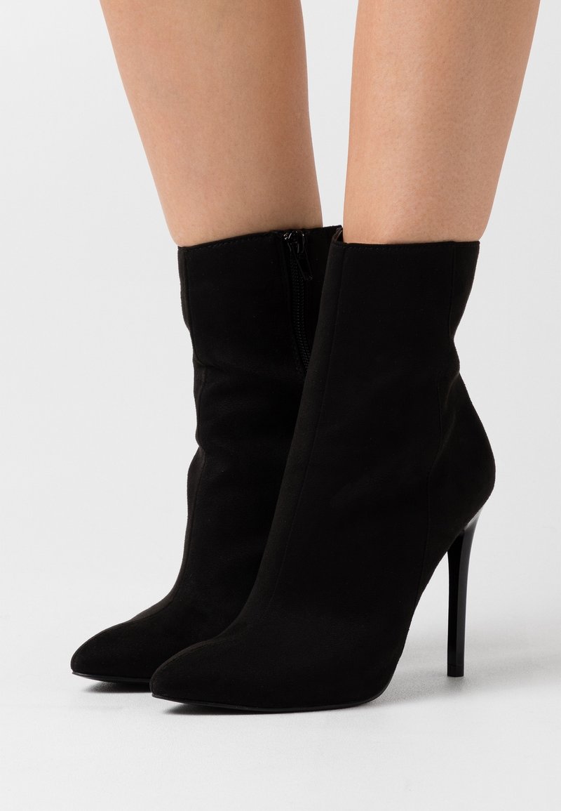 Femme portant des bottines en daim noir à bouts pointus et talons aiguilles, debout sur un sol blanc.
