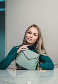 Mátově zelená kožená kabelka se strukturovaným tvarem, klopovým uzavřením, zlatými kovovými detaily a odnímatelným řetízkem, položená na mramorovém povrchu.
