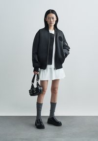 Μαύρο bomber μπουφάν με στρογγυλό πατς, λευκό μπλουζάκι, στρωμένο λευκό φόρεμα, γκρι καλτσοδέτες μέχρι το γόνατο και μαύρα ογκώδη παπούτσια, κρατώντας μια μικρή μαύρη τσάντα.