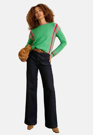 Pull vert avec des rayures roses et rouges, associé à un jean large bleu foncé et des bottines beige. Le modèle tient un sac beige.