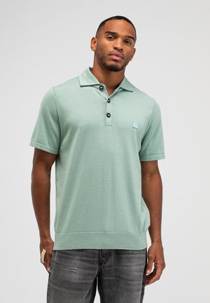 Man draagt een lichtgroen poloshirt met korte mouwen en knopen, gecombineerd met donkergrijze jeans met slijtage-effect, staand tegen een effen achtergrond.