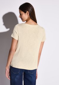 Top corto a maniche corte color beige chiaro con scollatura rotonda, caratterizzato da una consistenza morbida. Abbinato a pantaloni in denim blu scuro, che mostrano un look casual.